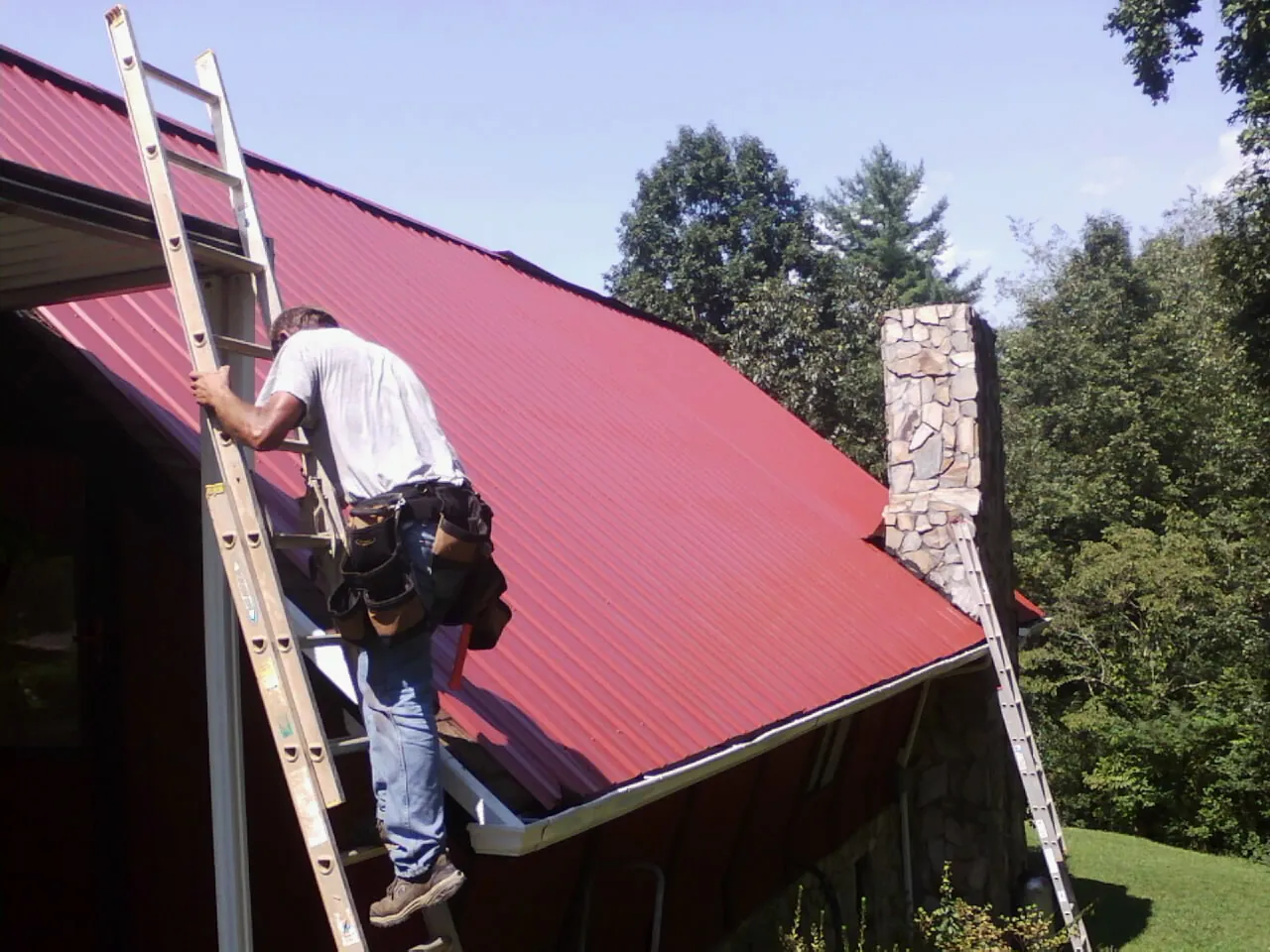 Roofer on a ladder beside a red metal roof and stone chimney — Soffit & Fascia Repair by Roofing Brothers in Annandale
